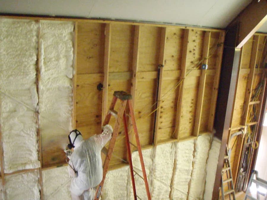 A technician in protective gear spraying closed-cell foam onto a framed wall