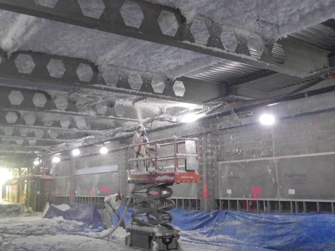 A technician on a lift spraying closed-cell foam onto the steel beams and metal deck of a large structure